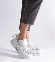 Model wearing white chunky sneakers with metallic accents, paired with ankle-length pants and a bracelet.