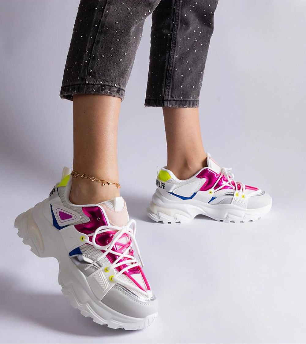 Woman wearing vibrant chunky sneakers with pink and neon accents, paired with black bejeweled jeans. Side view against a white backdrop.