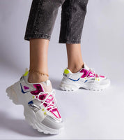Woman wearing vibrant chunky sneakers with pink and neon accents, paired with black bejeweled jeans. Side view against a white backdrop.