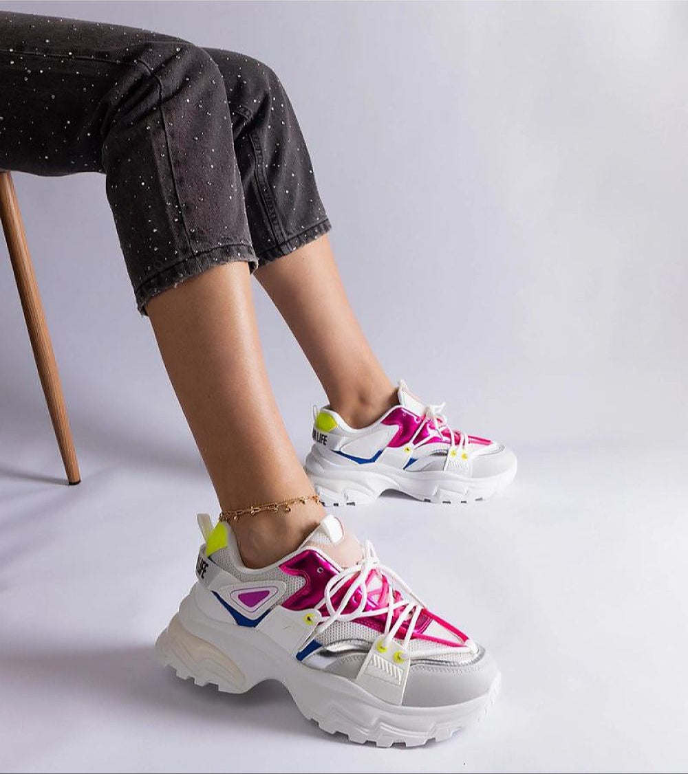 Colorful chunky sneakers with lace-up closure, worn by a seated person in black pants, featuring a bold and modern design.