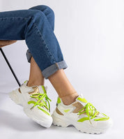 Person wearing colorful chunky sneakers with green accents and rolled-up jeans, sitting on a chair, showcasing modern sporty footwear style.