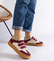 Person wearing stylish sneakers with burgundy and cream velcro design, brown sole, paired with rolled-up jeans on a wicker chair backdrop.