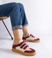 Person wearing maroon and white Velcro sneakers with gum soles, sitting on a chair in rolled-up jeans. Stylish and casual footwear.
