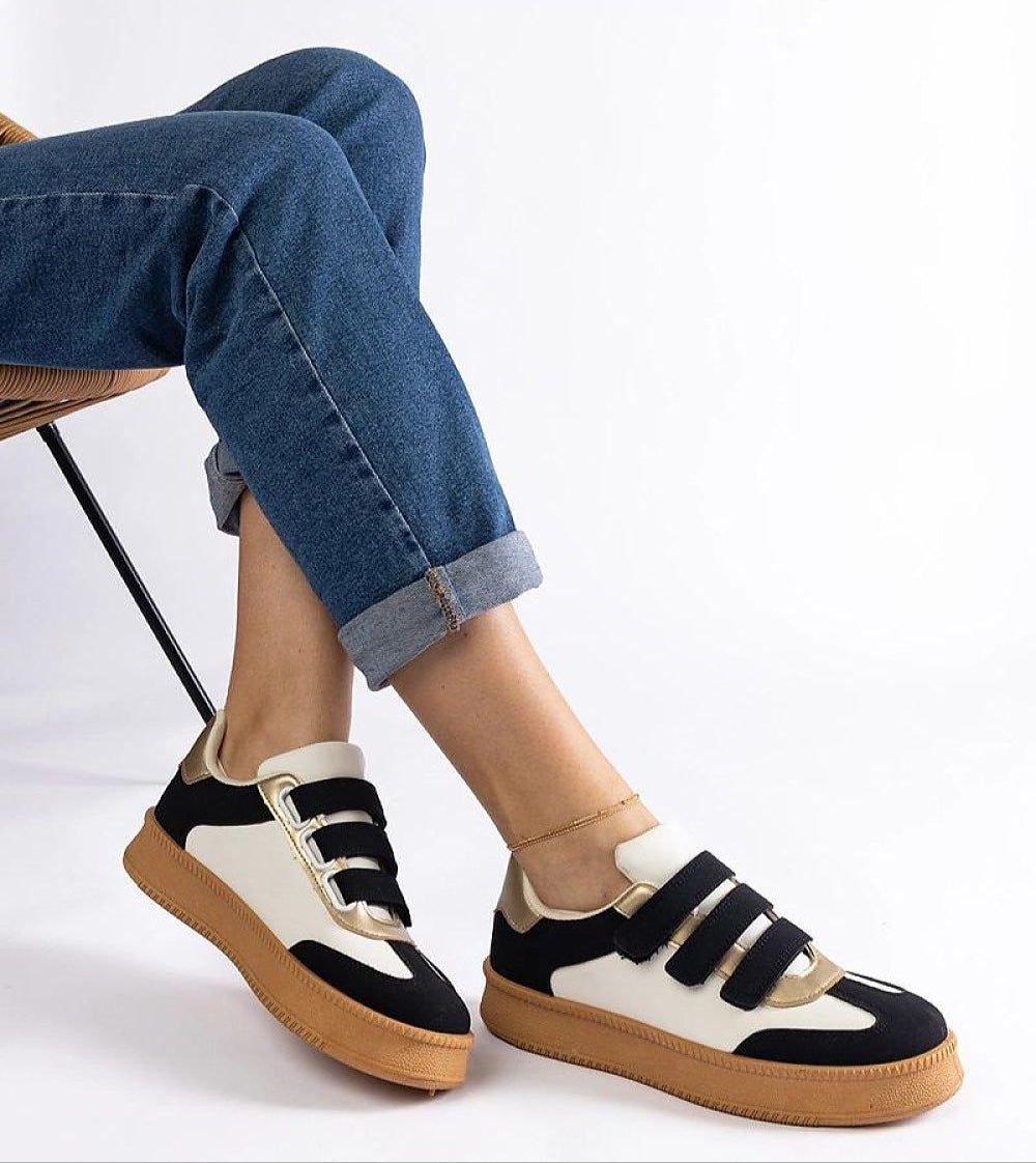 Person wearing Elurea Gold Velcro Street Sneakers with jeans, featuring golden sheen, eco leather, and brown rubber sole on a chair.