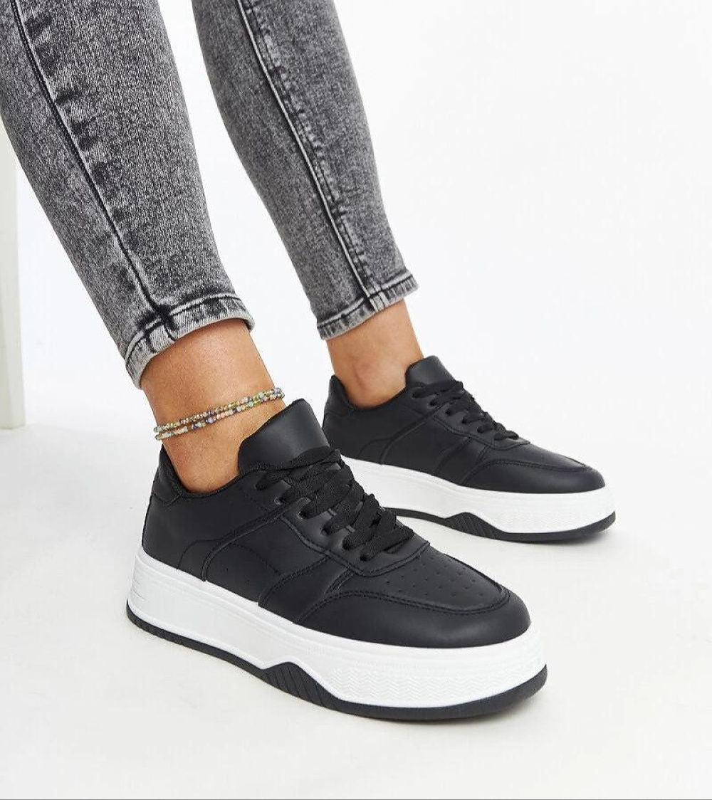 Woman wearing Kaedira Bold Platform Sneakers with black elevated sole and matte eco leather upper, styled with gray denim jeans.