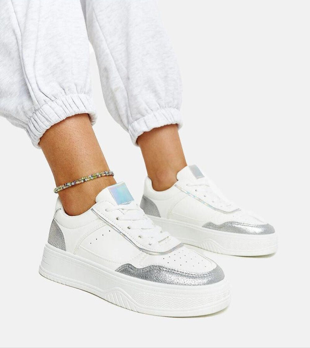 Person wearing Norevia silver-edged sneakers, featuring white platform sole and lace-up design with silver accents on a neutral background.