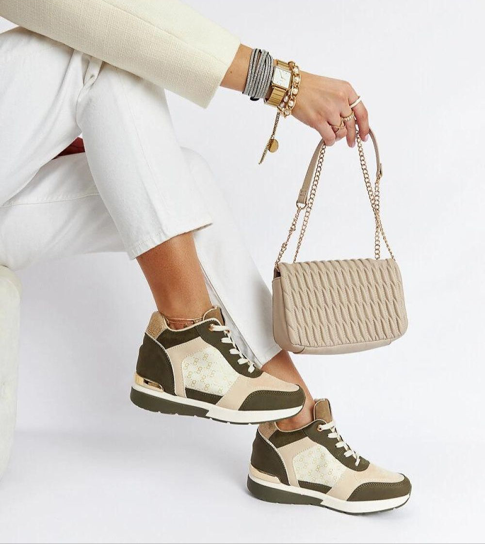 Stylish woman in beige outfit and accessories wearing olive wedge sneakers, holding quilted bag, showcasing casual elegance with lift.