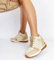 Stylish beige wedge sneakers with lace-up design and perforated details, paired with a tan handbag, worn by a woman seated.