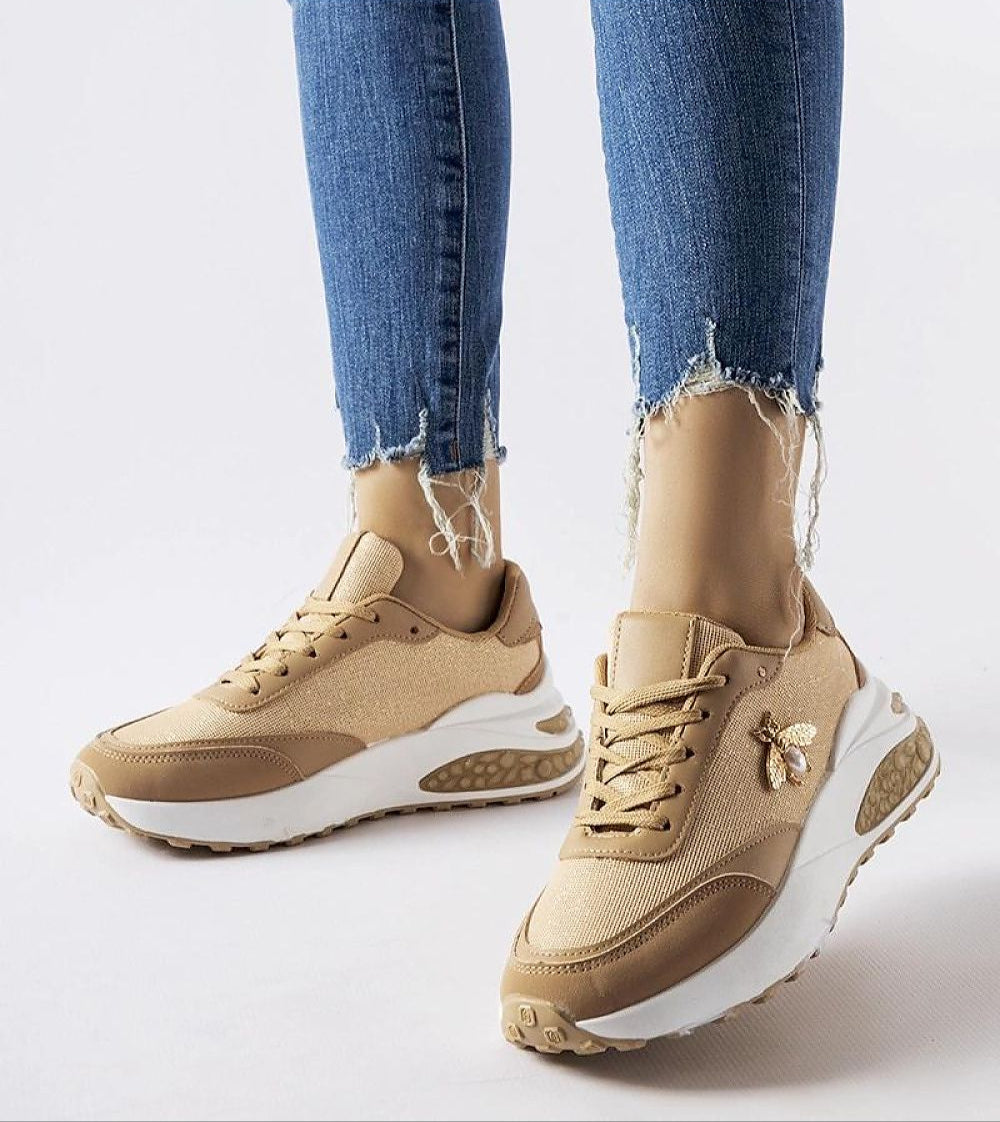 Woman wearing tan and white sneakers with embellishments, paired with distressed jeans, showing side view of stylish footwear.