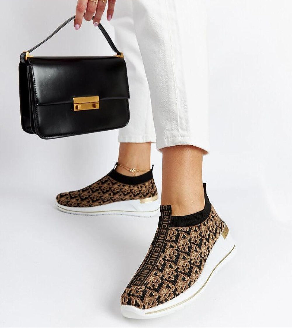 Woman wearing black-brown Marronza Slip-On Wedge Sneakers with white pants, holding a black handbag.