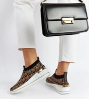 Woman wearing Marronza slip-on wedge sneakers and holding a black handbag. Sneakers have black-brown upper and two-tone rubber sole.