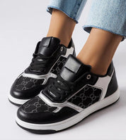 Black and white sneakers with monogram pattern and lace-up design, shown on feet wearing denim jeans.