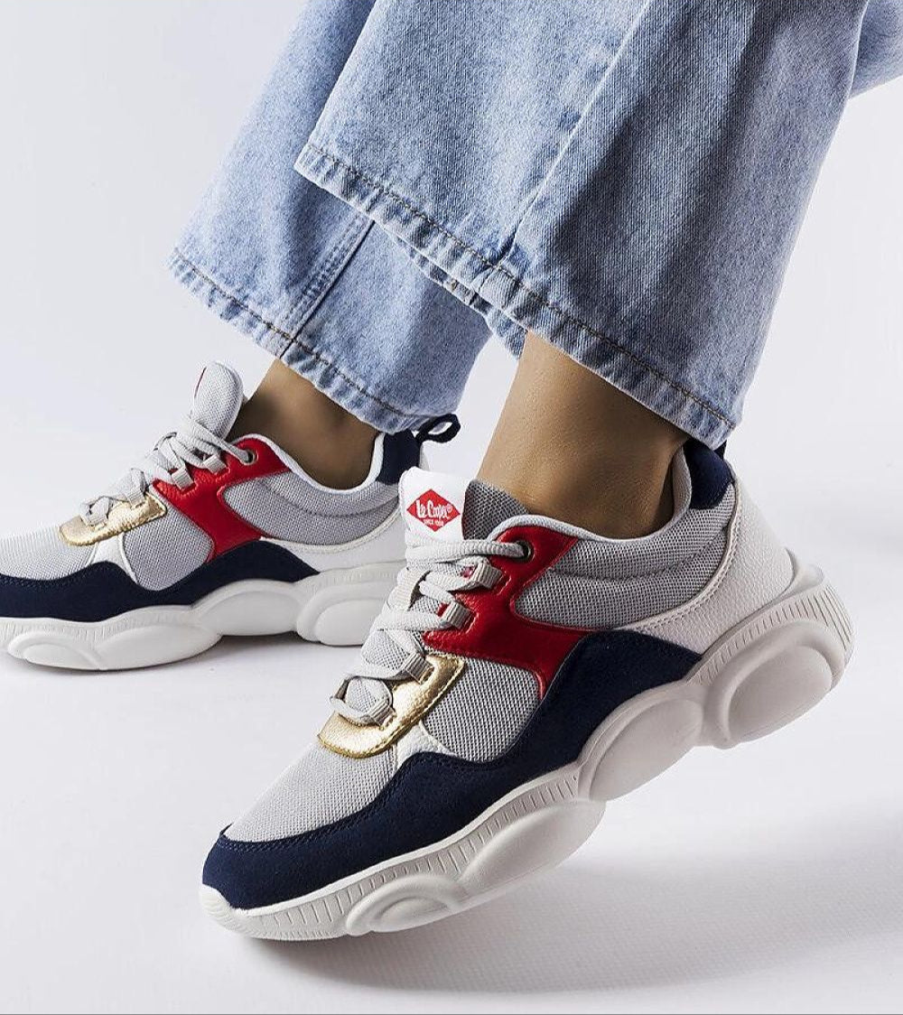 Person wearing gray sporty lace-up sneakers with red and navy accents, featuring a thick white sole, paired with blue jeans.