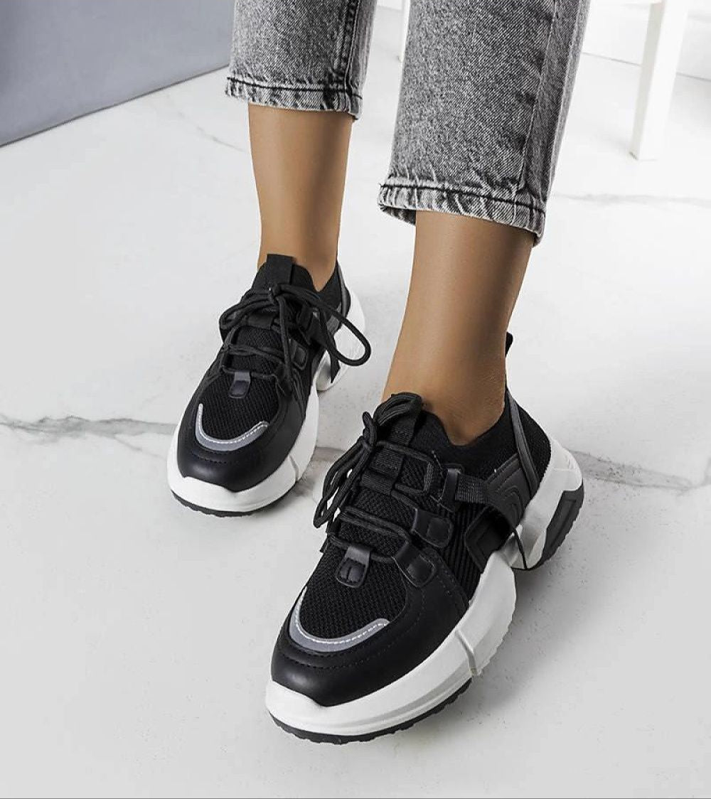 Black knit sneakers with white platform sole, showcasing their modern design and sock-like fit, worn with gray jeans on a marble floor.