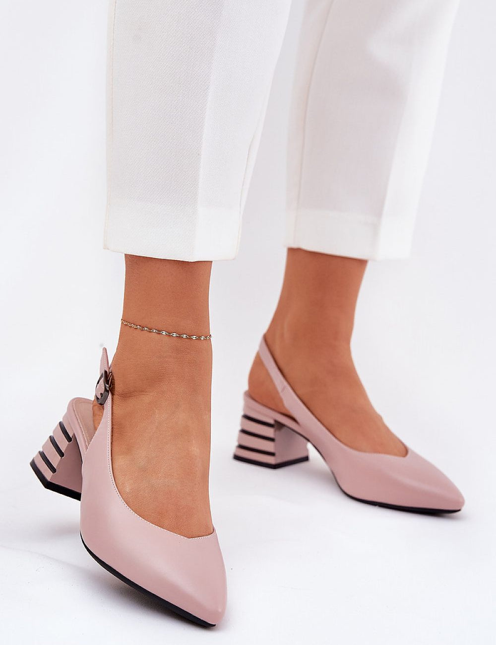 Woman wearing Elorelle Column-Heel Sandals in pink eco leather with ankle strap and pointed toe, paired with white trousers, day or evening wear.
