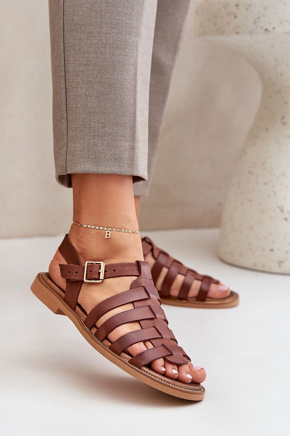 Woman wearing Celira Flat Leather Sandals with ankle buckle in brown, showcasing a sleek, elegant design perfect for versatile wear.