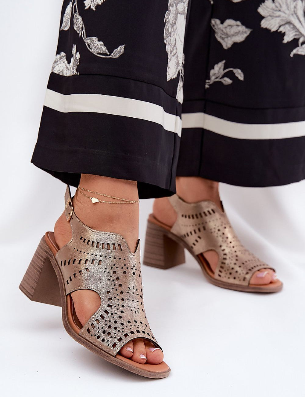 Woman wearing stylish cutout block heel sandals with ankle strap, accessorized with a delicate anklet, paired with floral print pants.