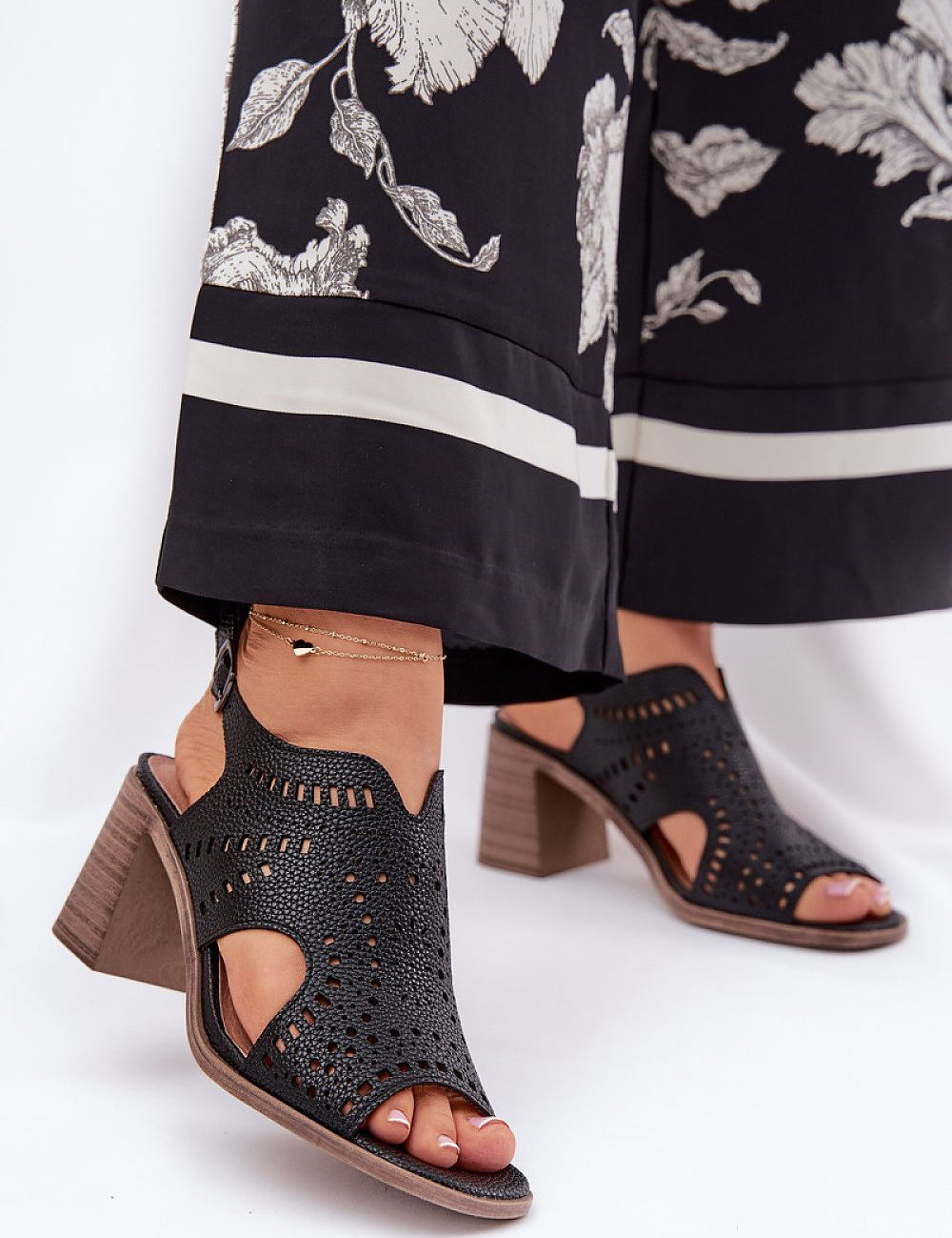 Black block heel sandals with cut-out design and ankle strap worn with floral-print black pants.