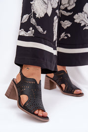 Black block heel sandals with cut-out design and ankle strap worn with floral-print black pants.