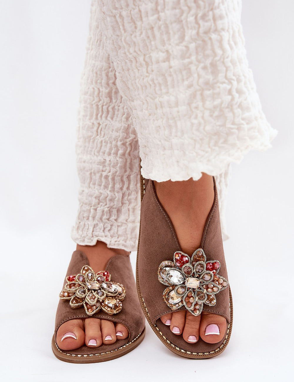 Woman wearing Fiorenna Flat Suede Flower Sandals with decorative floral accent and ankle straps, paired with white textured pants.