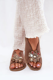 Woman wearing Fiorenna Flat Suede Flower Sandals with decorative floral accent and ankle straps, paired with white textured pants.