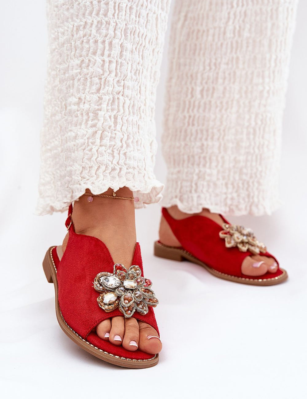Woman wearing Fiorenna Flat Suede Flower Sandals with sculpted flower accent and adjustable ankle strap, paired with white textured pants.