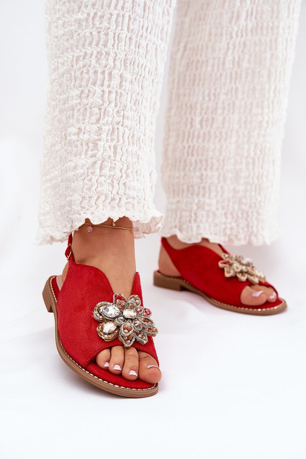 Woman wearing Fiorenna Flat Suede Flower Sandals with sculpted flower accent and adjustable ankle strap, paired with white textured pants.