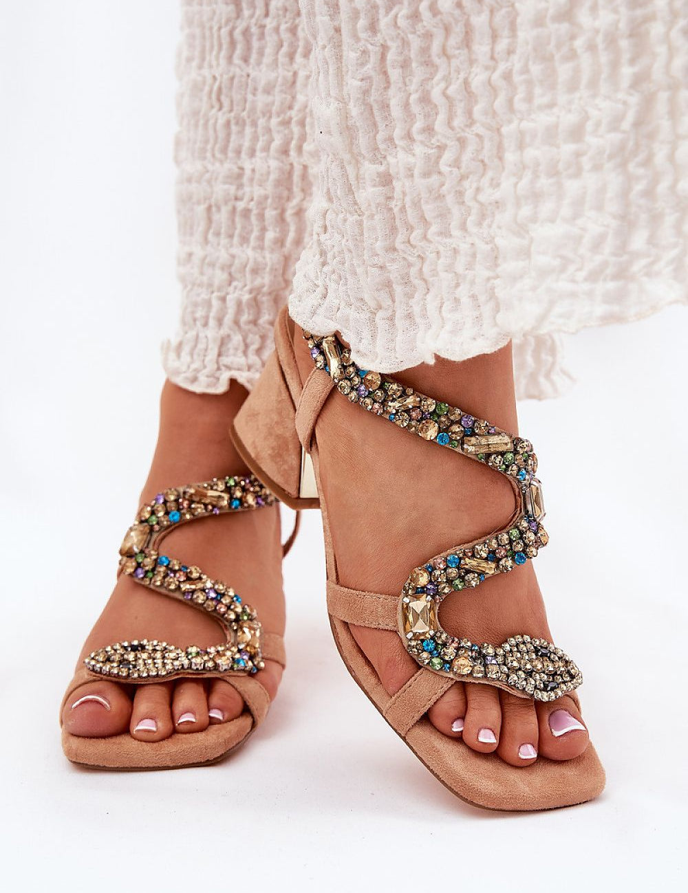 Beige serpent heel sandals with crystal accents on a woman's feet in a white dress, elegant footwear for formal occasions.