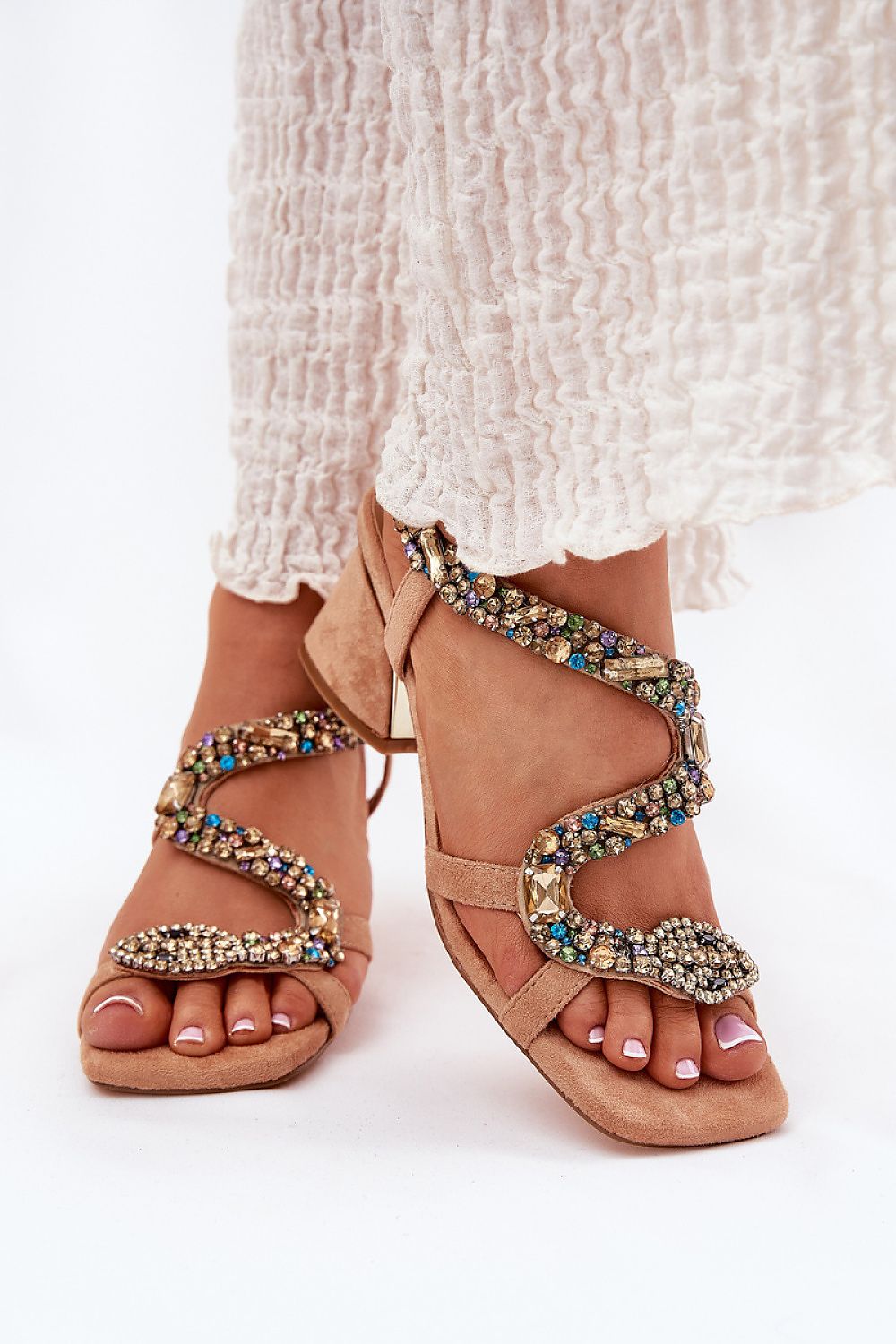 Beige serpent heel sandals with crystal accents on a woman's feet in a white dress, elegant footwear for formal occasions.