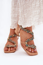 Beige serpent heel sandals with crystal accents on a woman's feet in a white dress, elegant footwear for formal occasions.