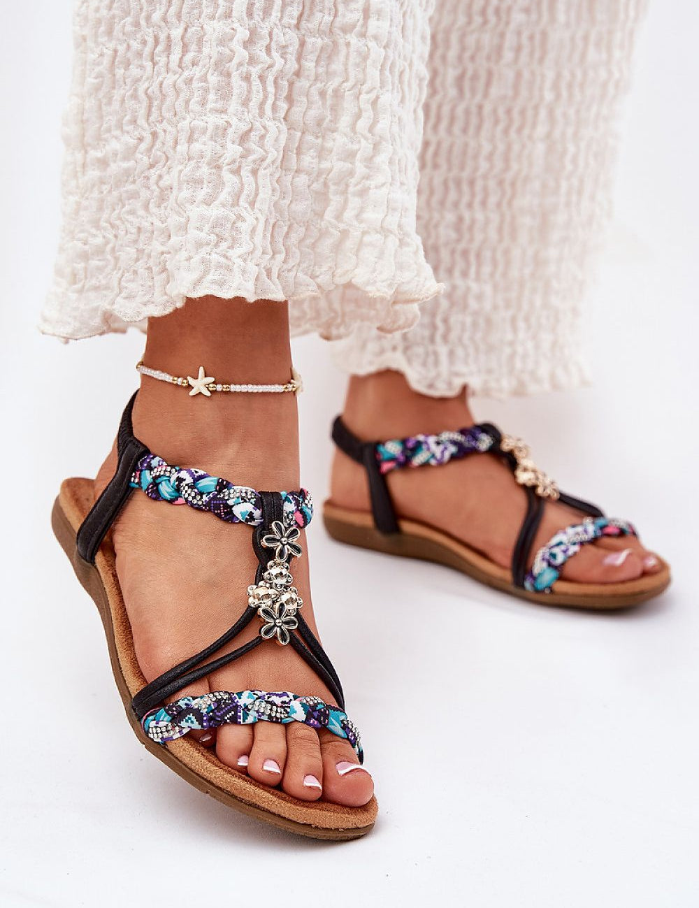 Close-up of Aviretta Braided Zircon Sandals with braided straps and zircon accents, worn with white textured pants.