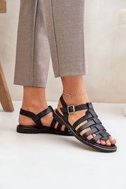 Woman wearing Celira Flat Leather Sandals in black with ankle buckle, styled with gray trousers, showcasing chic everyday elegance.