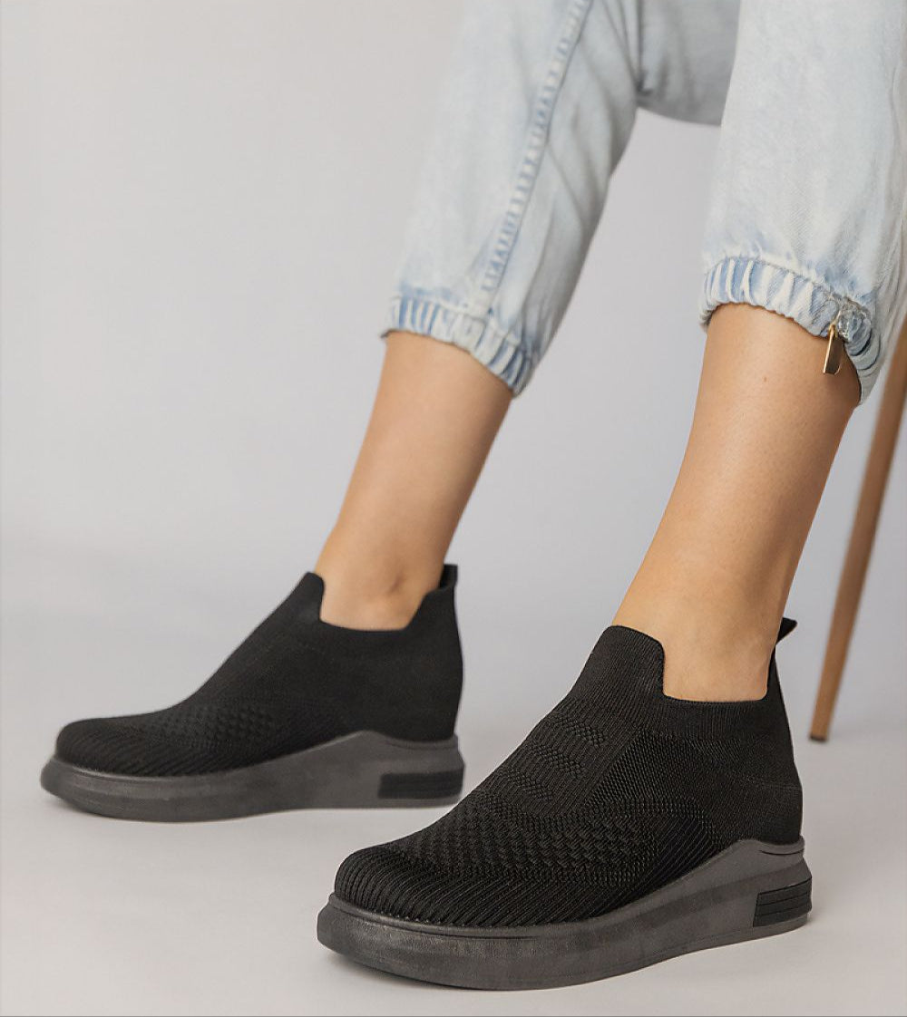 Black slip-on sock sneakers with stretch-knit upper and platform sole, worn with light denim jeans, showcasing minimalist urban style.