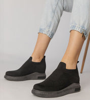 Black slip-on sock sneakers with stretch-knit upper and platform sole, worn with light denim jeans, showcasing minimalist urban style.