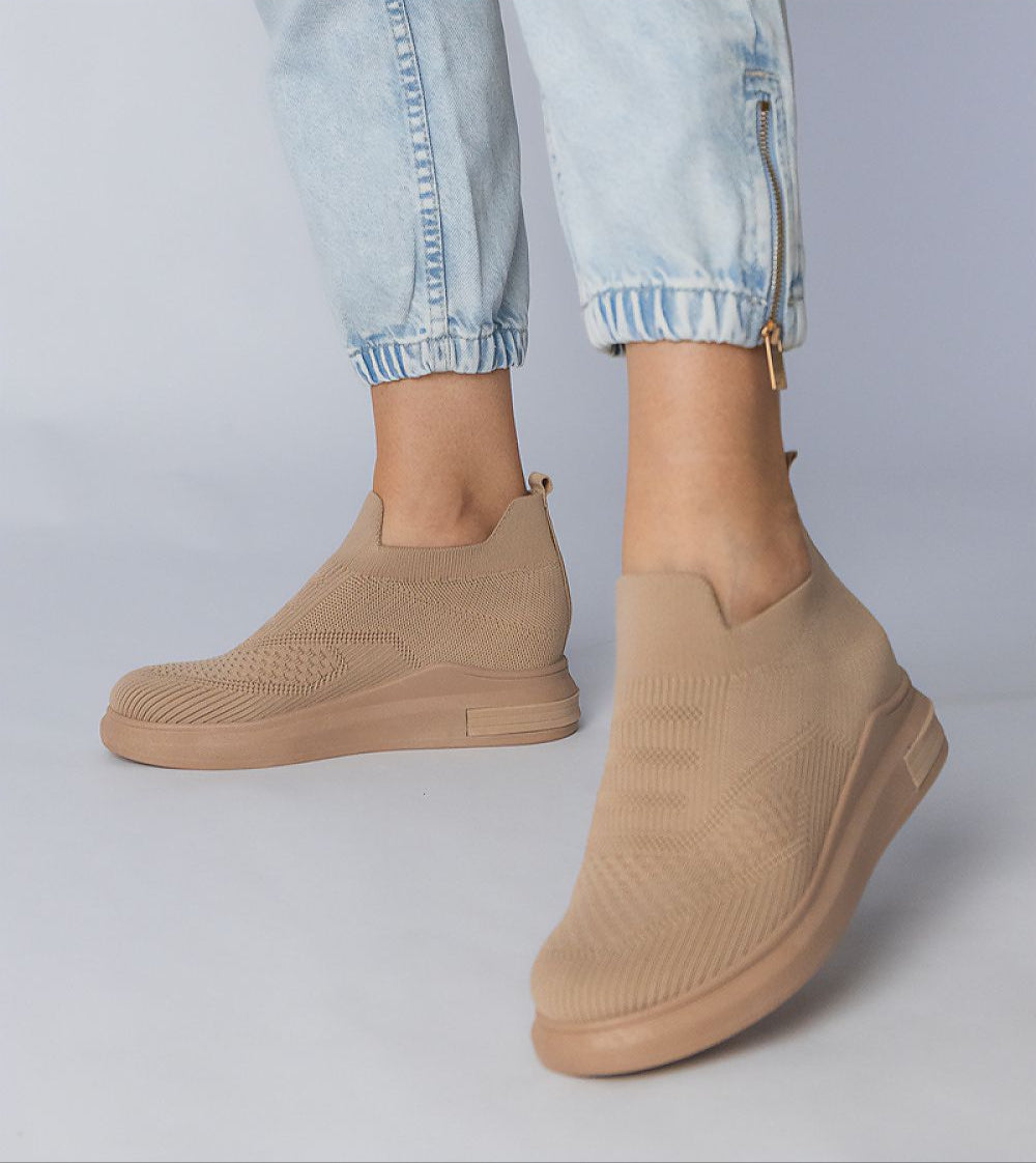 Beige slip-on sock sneakers with stretch-knit upper and platform sole, paired with light blue jeans for an urban chic look.
