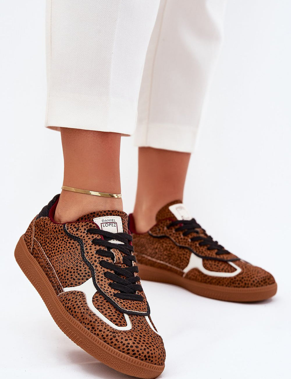 Close-up of a person wearing brown speckled sneakers with black laces and white accents, paired with white pants.