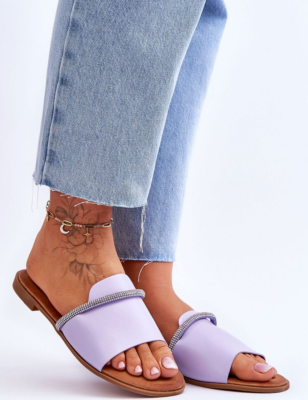 Purple slip-on sandals with rhinestone straps worn by person in blue jeans, displaying a stylish and casual summer footwear choice.
