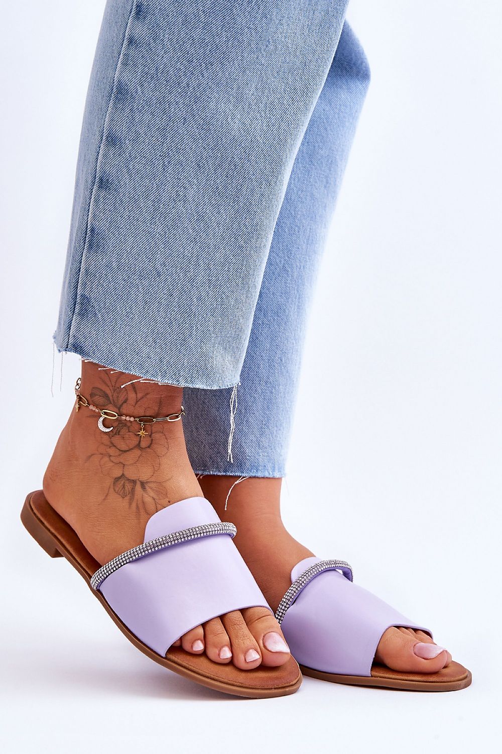 Purple slip-on sandals with rhinestone straps worn by person in blue jeans, displaying a stylish and casual summer footwear choice.