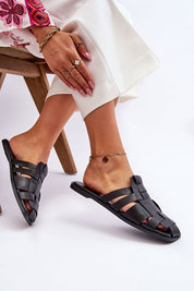 Woman wearing Rovella slip-on flat flip-flops in black eco leather with a low-profile heel, seated casually on a wooden stool.
