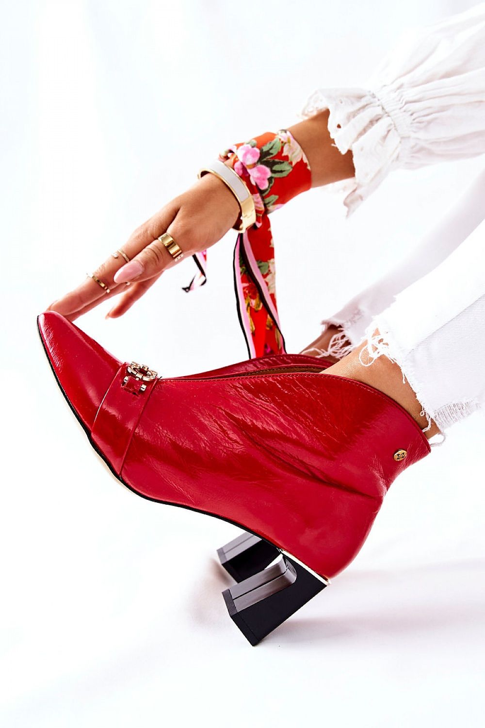 Red leather high heel boot with decorative accents, paired with white jeans and floral bracelet, showcasing elegant and modern fashion style.