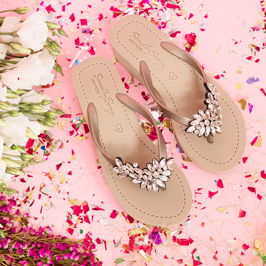 Elegant rhinestone sandals on pink confetti background with white and purple flowers, perfect for summer fashion.