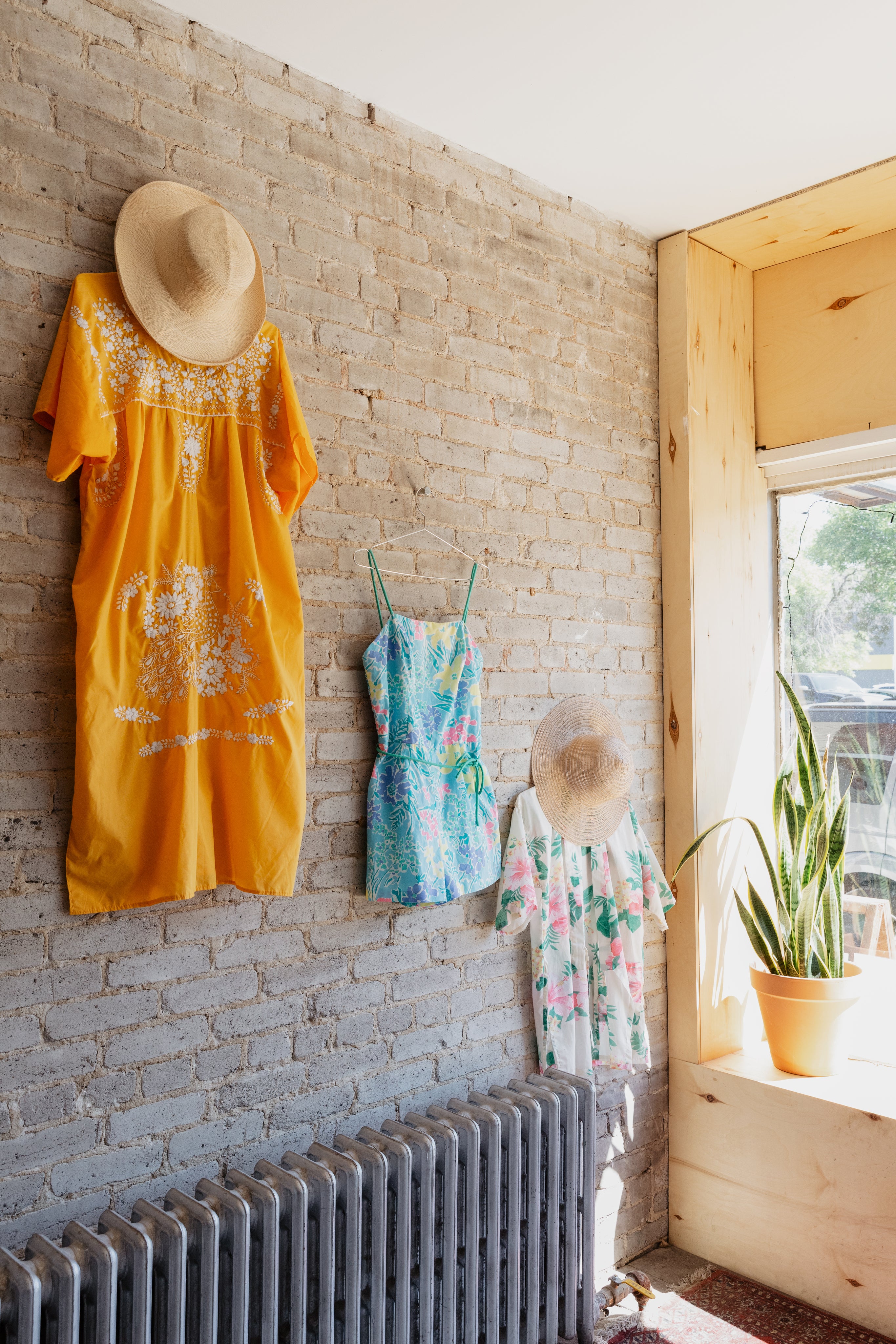 Three summery dresses and hats on a rustic brick wall near a sunny window with a potted plant nearby.
