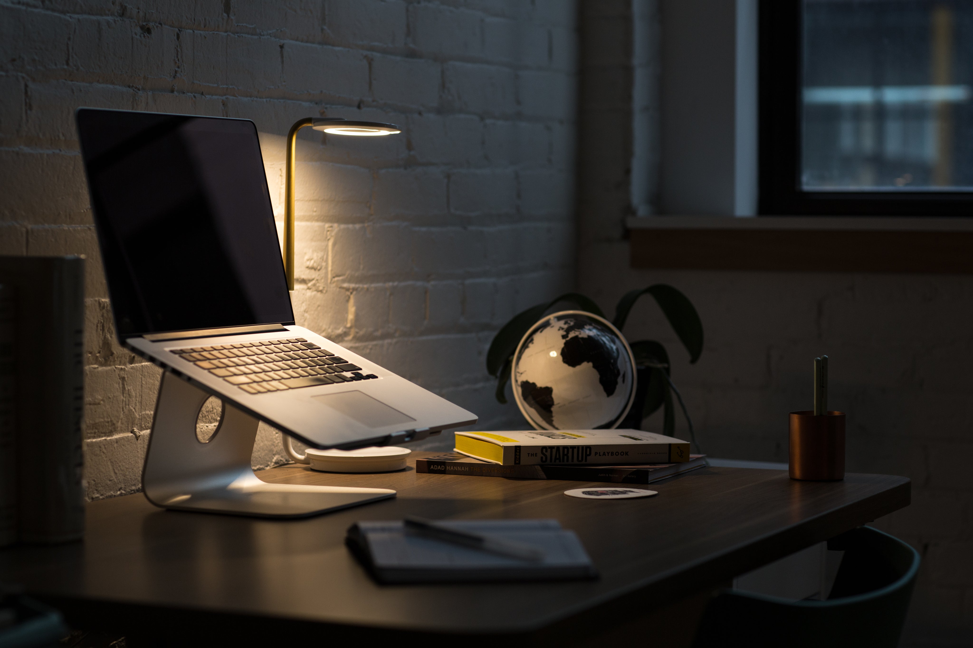 Stylish home office setup with a laptop, desk lamp, globe, and books on a wooden table against a brick wall.
